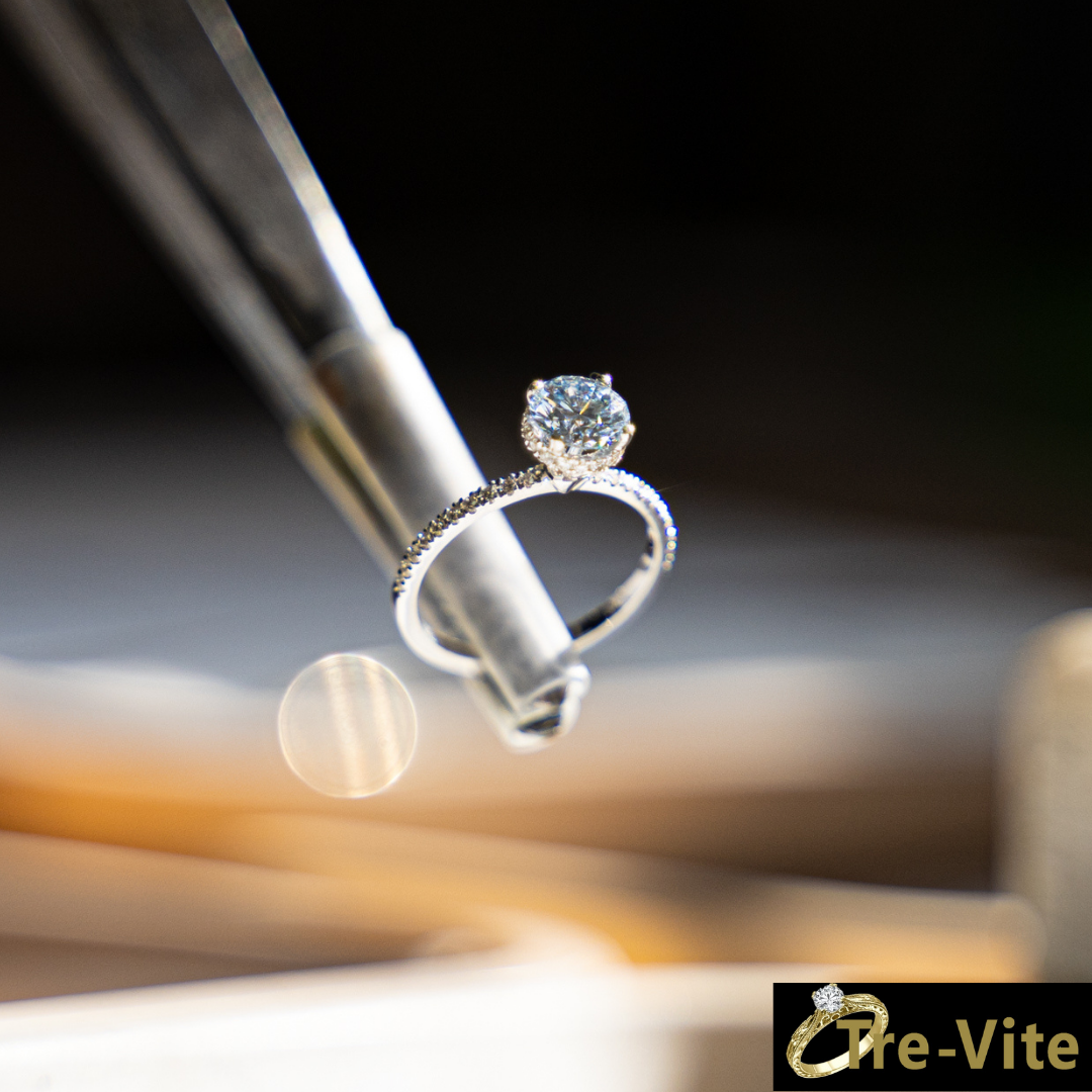 טבעת יהלום אלגנטית מוחזקת באמצעות פינצטה מתכתית. היהלום המרכזי בצורת עגולה, מוקף בהילה של יהלומים קטנים, והחישוק עצמו משובץ ביהלומים זעירים. הרקע מטושטש ומואר באור רך, מה שמדגיש את הברק של היהלום. בפינה הימנית התחתונה מופיע לוגו של המותג 
