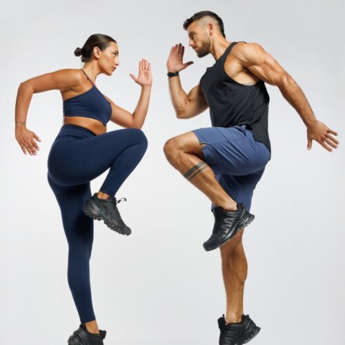 בתמונה, על רקע בהיר חלק, עומדים שני ספורטאים – אישה משמאל וגבר מימין – בבגדי אימון וצעדי תרגיל “הרמת ברך גבוהה” (high knees). האישה לובשת חזיית ספורט וגרביון בגוון כחול כהה, נעלי ספורט לבנות, ושערה אסוף לאחור. היא מרימה את ברך ידה הימנית לגובה חזה, זרועותיה כפופות בזווית של כ־90° ומניחות בפנים. הגבר לובש גופיית ספורט שחורה ומכנסיים קצרים בגוון כחול, נעלי ספורט שחורות, ושערו גזור קצר. הוא מרימה את ברך רגלו השמאלית עד לגובה דומה, ידיו כפופות ומונחות מבעד לגופו באותה זווית. שניהם ניצבים בהתכופפות קלה, מביטים זה על זו, ומייצרים תחושה של תרגול זוגי או תחרות ידידותית.